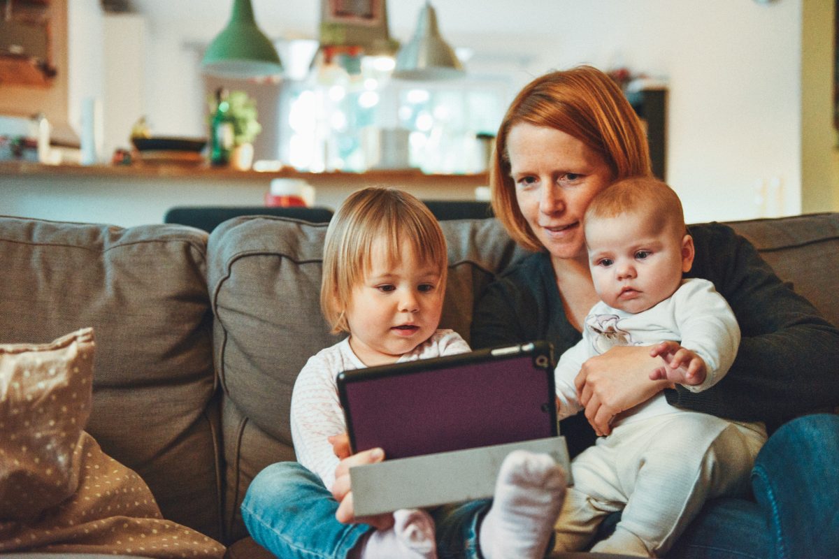 Eine Mutter schaut mit ihrem Baby und ihrem Kleinkind etwas auf dem Tablet an.