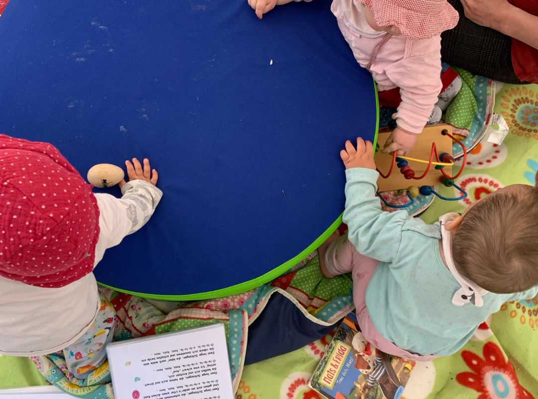 Das Bild zeigt eine Impression von oben auf eine Spielsituation für Kleinkinder. Es sind deri Kleinkinder, Spielzeuge, Liedtexte zu sehen.