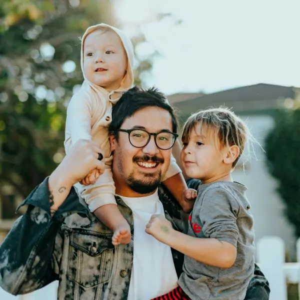 Ein lächelnder Papa mit zwei Kindern, das eine Kind sitzt auf seine Schulter, das andere Kind auf seinem Arm