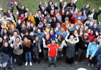 Ein Gruppenfoto der Mitarbeitenden des Mütterzentrum e.V. Leipzig, aufgenommen aus der Vogelperspektive: Personen unterschiedlichen Alters Lachen und Winken in die Kamera.