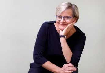 Eine Portraitaufnahme der Hebamme Raja Rohrwacher, die freundlich in die Kamera lächelt. Sie trägt kurzes graues Haar, ein dunkles Kleid und eine Brille mit dunklem Rahmen.