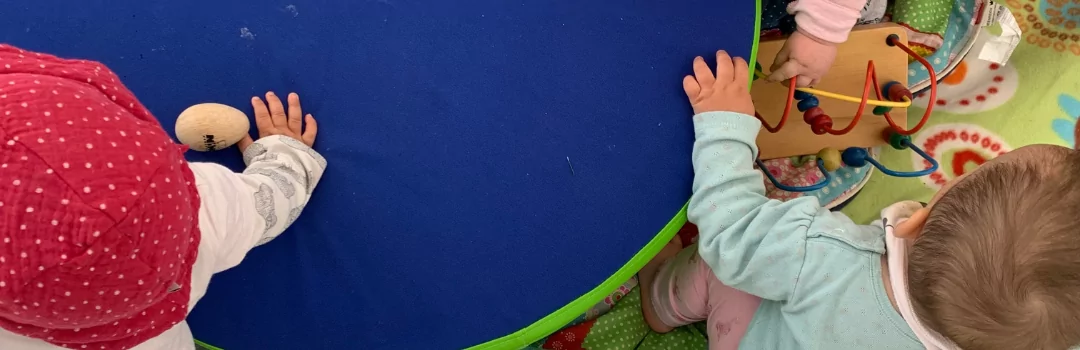 Das Bild zeigt eine Impression von oben auf eine Spielsituation für Kleinkinder. Es sind deri Kleinkinder, Spielzeuge, Liedtexte zu sehen.
